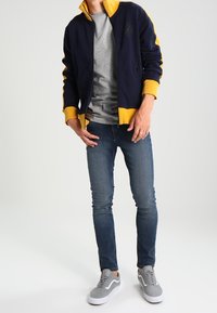 Marinblå idrottsjacka med gula detaljer, grå t-shirt, medelblå jeans och grå sneakers med vita sulor.