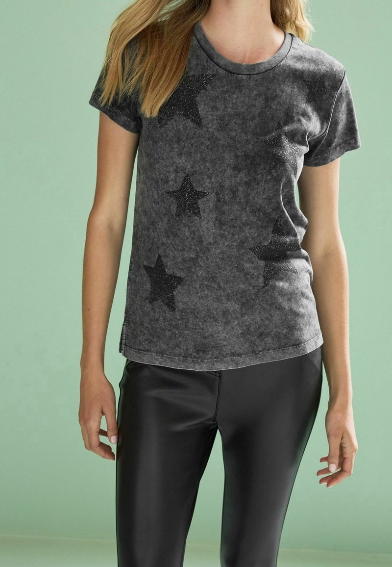 Grå t-shirt med et falmet udseende, prydet med sorte glimmerstjerner. Korte ærmer og rund halsudskæring, parret med sorte leggings.