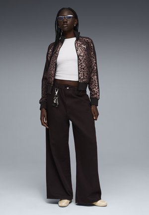 Vrouw met gevlochten haar, blauwgetinte bril, leopardprint Puma-jas, wit cropped topje, wijde zwarte broek met sleutelhanger en beige schoenen.