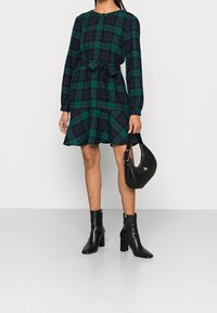 Robe à carreaux verts et bleu marine avec manches longues, fermeture à boutons, et ceinture à la taille. Associée à des bottines noires à talons et un petit sac à main noir.