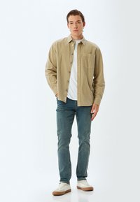 Camicia button-up beige chiaro sopra una t-shirt bianca a girocollo, abbinata a jeans blu e sneakers bianche con dettagli beige.