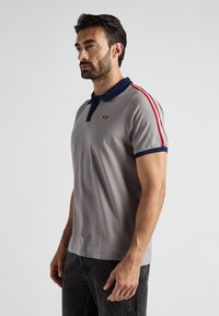 Szary polar z granatowym kołnierzykiem i czerwonymi paskami po bokach. Posiada logo na klatce piersiowej oraz krótkie rękawy. Gładka struktura materiału.