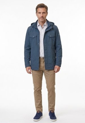 Hombre con chaqueta azul con capucha, camisa estampada, pantalones beige y zapatillas azules, de pie mirando hacia adelante sobre fondo blanco.