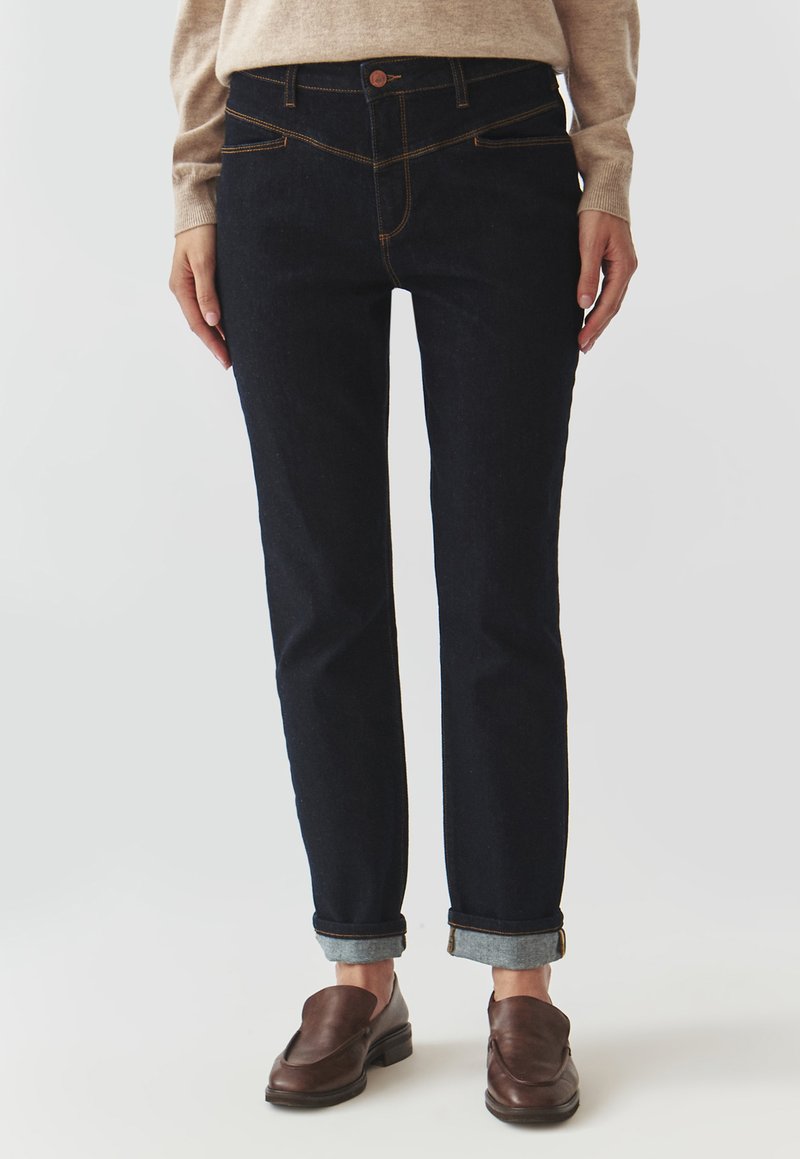 Jeans din denim închis, cu o croială slim, design cu cinci buzunare, manșete rulouri și cusături contrastante. Purtați cu un pulover tricotat bej și pantofi maro.