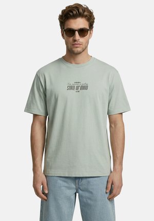 Man met zonnebril, lichtgroen t-shirt met tekst "STATE OF MIND" en lichtblauwe spijkerbroek, staand tegen een effen achtergrond.