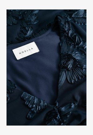 Vêtement bleu marine avec des appliqués floraux texturés, présentant un décolleté en V. Étiquette marquée "NOELLA" visible au centre. Fond en tissu lisse.