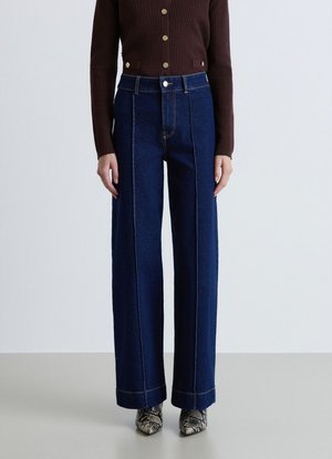 Donna che indossa jeans blu scuro a vita alta e gamba larga, maglione marrone con bottoni e stivaletti alla caviglia con punta a fantasia pelle di serpente, in piedi su pavimento grigio.