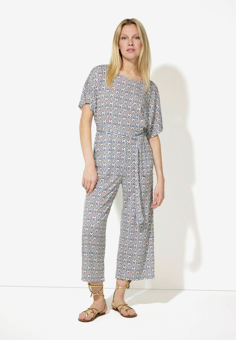 Gemusterter Jumpsuit in Blau, Weiß und Orange; kurze Ärmel, lockere Passform, taillierter Gürtel, weite Beine, kombiniert mit goldenen Sandalen mit Riemchen.