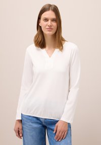 Weißes Langarm-V-Shirt aus weichem Stoff mit dezentem Texturmuster. Kombiniert mit hellblauen Jeans, was einen lässigen Look ergibt.