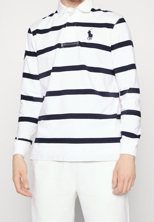 Homme portant un maillot de rugby blanc à manches longues avec de larges rayures horizontales noires et un logo de joueur de polo sur la poitrine, associé à un pantalon blanc.