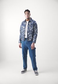 Chaqueta azul con estampado floral, camiseta blanca, jeans azules y zapatillas grises. La chaqueta tiene un cierre de cremallera y una textura sutil.
