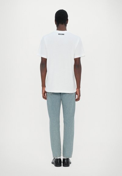 T-shirt blanc à manches courtes avec un logo "Moschino" noir au dos, associé à un jean droit bleu clair et des chaussures noires.