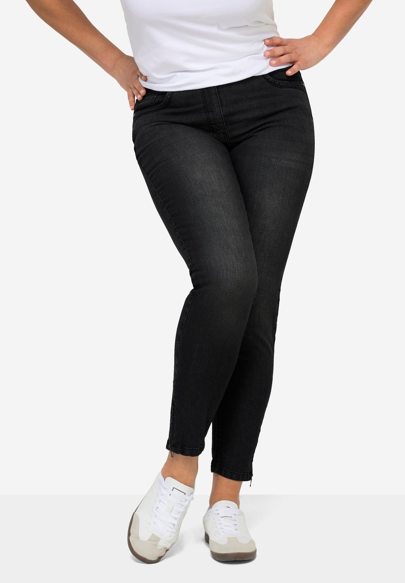 Persona che indossa jeans neri aderenti e sneakers bianche, in piedi con le mani sui fianchi su uno sfondo semplice.