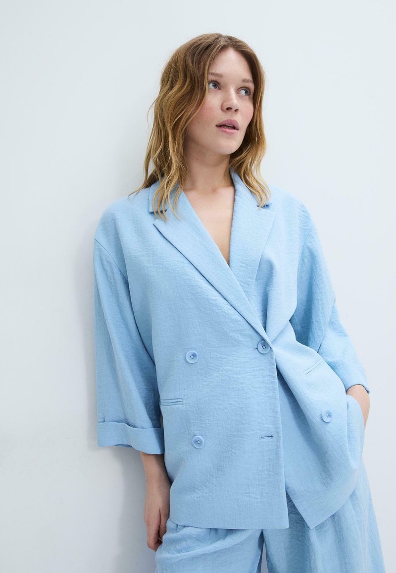 Femme aux cheveux ondulés châtain clair portant un blazer bleu clair ample à double boutonnage et un pantalon assorti, debout contre un mur blanc.
