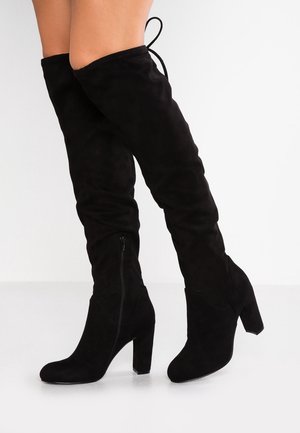 Knielange schwarze Wildlederstiefel mit einem klobigen Absatz, seitlichem Reißverschluss und Bindung oben. Die glatte Textur und das enganliegende Design betonen das Bein.