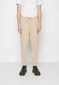 Beige Jogginghosen mit einem elastischen Bund und Kordelzug, mit schmal zulaufenden Beinen und gerippten Bündchen, getragen mit schwarzen Schnürstiefeln.