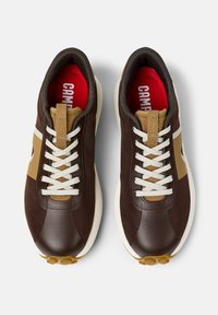 Brown sneakers with a textured fabric upper, tan accents, white laces, and red insoles featuring a logo. Thick rubber outsole with circular grip.