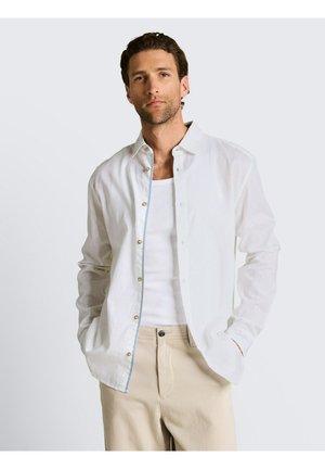 Mann trägt ein weißes Hemd mit Knöpfen über einem weißen Tanktop, beige Hose, steht mit den Händen in den Taschen vor einem schlichten Hintergrund.