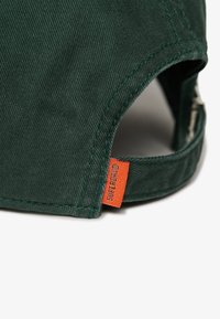 Gorra de algodón verde con visera curva, tela texturizada y una etiqueta naranja en el lateral. Detalle de costura visible a lo largo de los bordes.