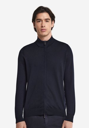 Homme aux cheveux foncés portant un pull zippé bleu marine et un pantalon foncé, debout devant un fond blanc uni.