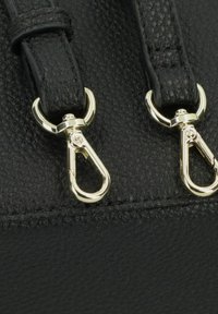 Nahaufnahme einer schwarzen, strukturierten Lederhandtasche mit zwei goldfarbene Metallclips, die an schwarzen Trageriemen befestigt sind und deren sichere Befestigung betonen.