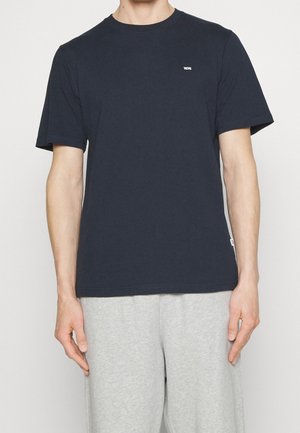 Mand iført en ensfarvet marineblå kortærmet t-shirt med lille hvidt logo på brystet og lysegrå joggingbukser, stående mod en hvid baggrund.