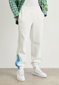 Hvide sweatpants med grønne bånd og blå detaljer, der har en elastisk talje og manchetter, parret med hvide sneakers på en neutral gulv.