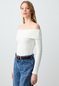 Weißes, schulterfreies, geripptes Langarmoberteil, kombiniert mit hoch taillierten blauen Jeans und einem braunen Gürtel mit strukturiertem Finish.
