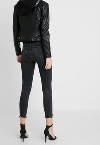 Zwarte leren jas, skinny donkergrijze jeans met rafelige zoom en zwarte hoge hakken. Zicht van achteren, met een getailleerde silhouet.