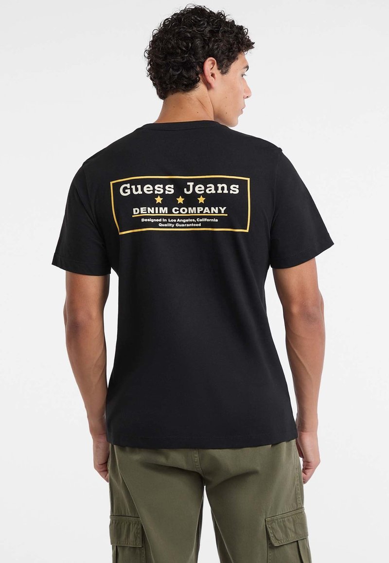 Mężczyzna w czarnej koszulce z napisem "Guess Jeans Denim Company" na plecach, w połączeniu z oliwkowymi spodniami typu cargo, odwrócony plecami.