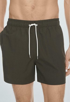 Torso e gambe maschili che indossano pantaloncini a vita elastica verde scuro con coulisse bianca, mani appoggiate ai lati, sfondo chiaro semplice.