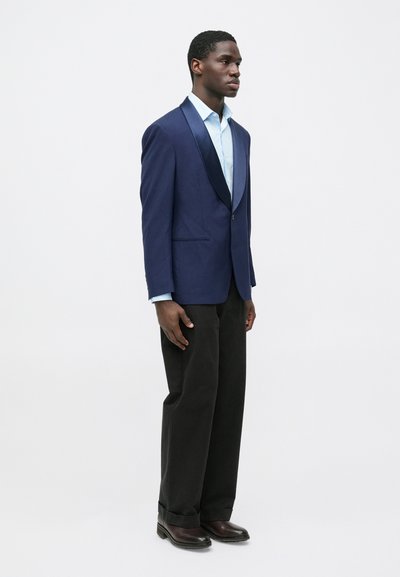 Tmavomodré sako s lesklým saténovým klopou, kombinované so svetlomodrou košeľou, tmavými nohavicami a hnedými topánkami; hladká látka a priliehavý strih.