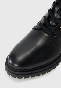 Bota de cuero negro con punta redondeada, superficie texturizada y cordones negros. Suela de goma sólida con dibujo para tracción. Ojales en tono dorado.