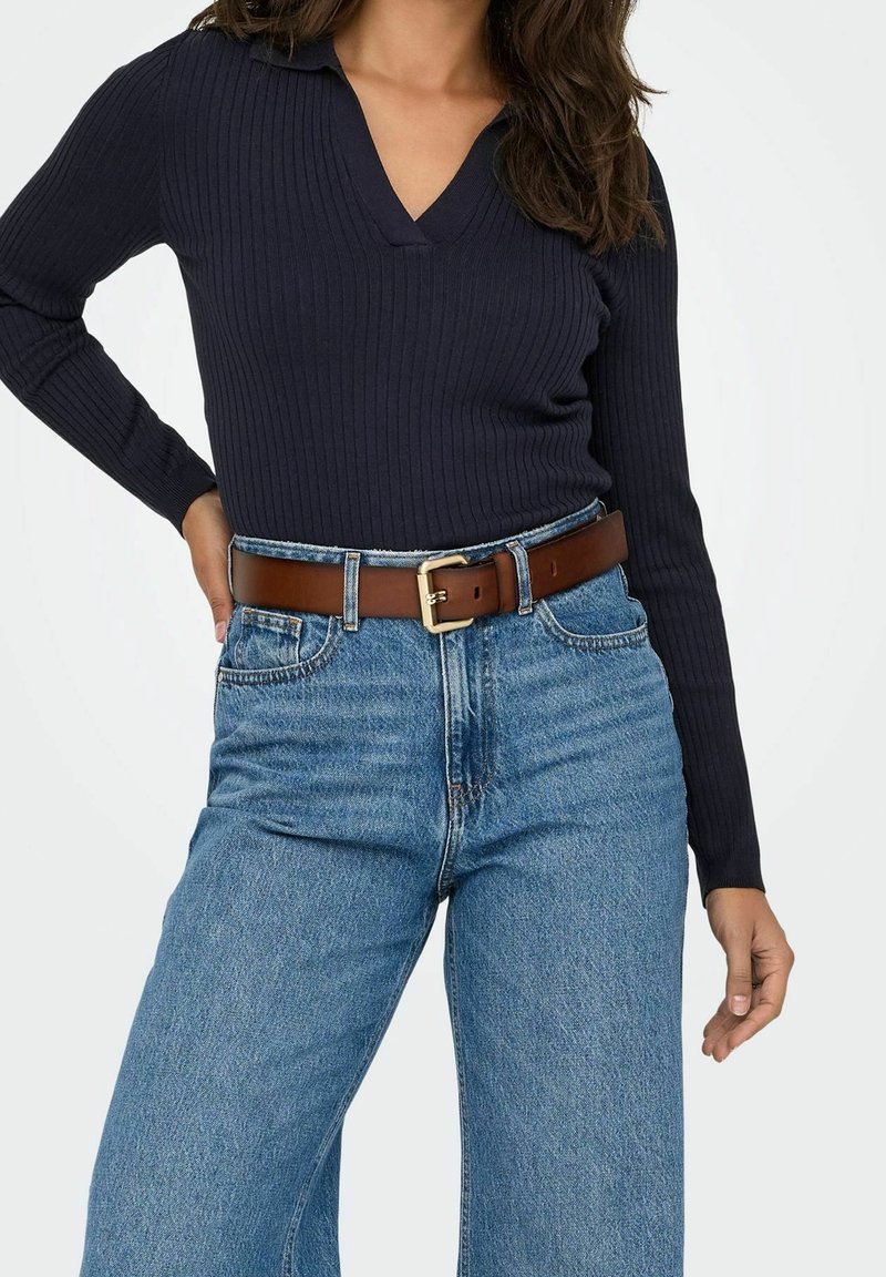 Femme portant un haut côtelé bleu marine à manches longues, rentré dans un jean bleu taille haute, accessoirisé d'une ceinture en cuir marron avec une boucle dorée.