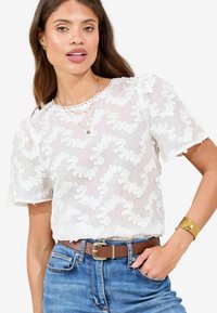 Witte kanten blouse met patroon, korte mouwen en ronde halslijn. Gecombineerd met hooggeplaats blauwe jeans en een bruine riem met een gouden gesp.