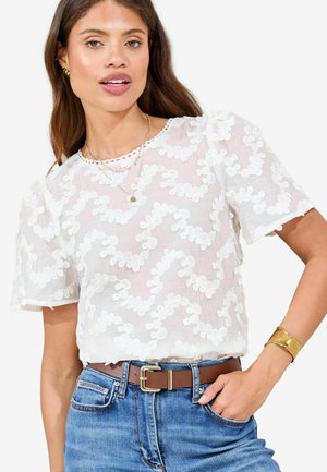 White lace blouse with pattern, short sleeves, and round neckline. Paired with high-waisted blue jeans and a brown belt with a gold buckle.