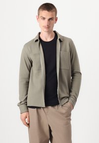 Svijetlo maslinasto zeleni zip-up sako s dva džepa na prsima, nošen preko crne majice, uparen s bež hlačama. Glatka tekstura i ležeran dizajn.