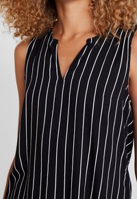 Svart ärmlös topp med vita vertikala ränder, V-ringning och lös passform, tillverkad av lättviktigt material. Ingen synlig hårdvara.