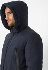 Uomo con barba corta che indossa una giacca a vento blu navy con cappuccio, chiusa con cerniera nera visibile e tasca laterale con cerniera, che guarda a sinistra.