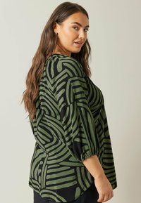 Blusa verde e nera con motivo a fantasia, dal taglio morbido, con maniche a palloncino e un design di linee astratte su un tessuto leggero.