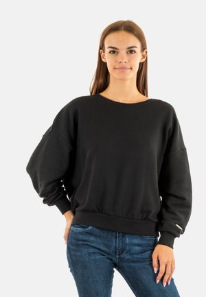 Sudadera negra hecha de una tela suave, con cuello redondo, mangas oversize y cintura ajustada. Combinada con jeans de mezclilla azules.