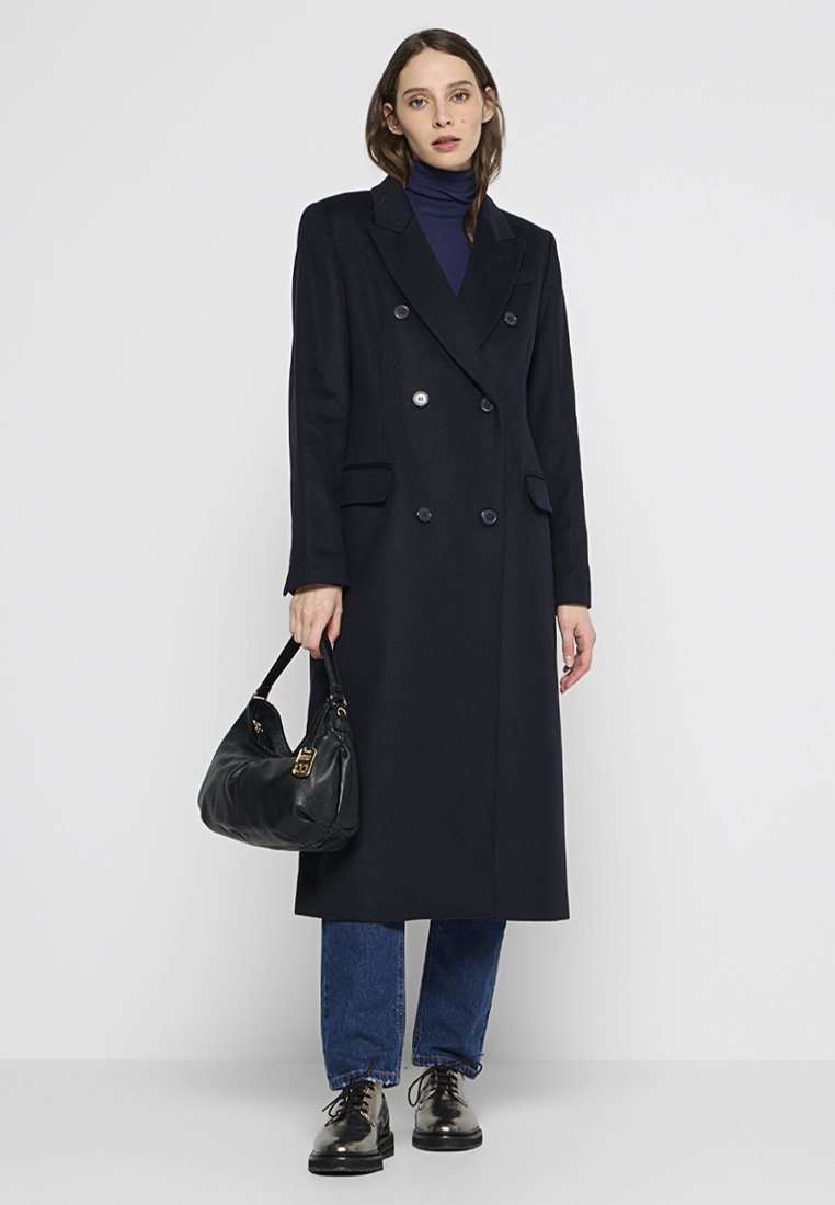 Marineblauwe wollen dubbele-breasted jas met knoopdetails, notch lapel en zijzakken, gecombineerd met een zwarte tas en donkere jeans.