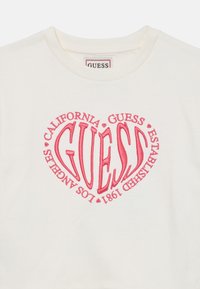 Sweatshirt en coton blanc présentant un grand graphique de cœur rose avec "GUESS" et du texte environnant. Encolure ronde et texture douce.