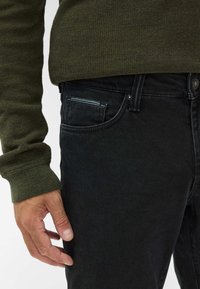 Jeans in denim nero con una vestibilità aderente, dotati di una tasca laterale con cuciture a contrasto. Un maglione verde testurizzato è visibile sopra.