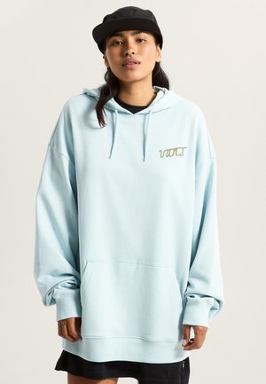 Hellblauer, übergroßer Hoodie mit Fronttasche, gerippten Bündchen und einem goldbestickten Logo auf der Brust. Schwarze Mütze getragen.