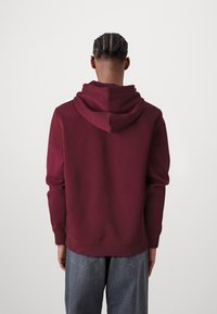 Felpa con cappuccio bordeaux dal tocco morbido, polsini a coste e vestibilità rilassata. Indossata con jeans baggy blu chiaro su uno sfondo neutro.