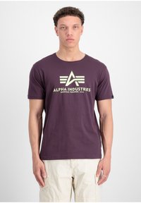 Lila Baumwoll-T-Shirt mit kurzen Ärmeln, das ein großes weißes Logo und ein grafisches Design auf der Vorderseite zeigt. Einfacher Rundhalsausschnitt.