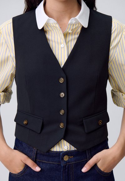 Una persona indossa un gilet nero sopra una camicia a righe gialle e bianche, con le mani appoggiate sulle tasche di jeans scuri. Sfondo neutro.