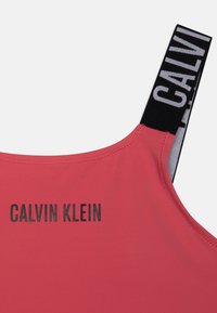 Roza kratka majica s tankimi črnimi naramnicami, ki ima belo oznako z logotipom in črn napis "CALVIN KLEIN" na hrbtni strani. Gladka tekstura tkanine.