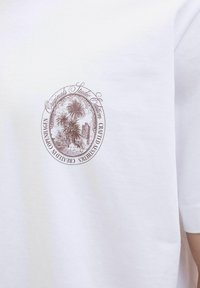 Hvid bomulds T-shirt med et cirkulært brunt grafik af palmer og arkitektur, med tekst der beskriver studio edition og håndværk.
